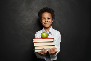 Happy black child student holding books and apple and smiling on chalkboard background