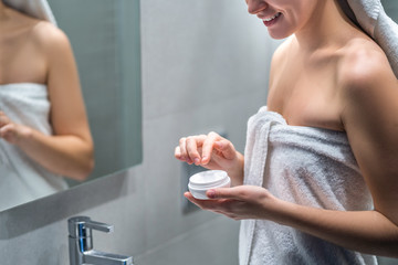 Happy woman in a bath towel holds moisturizing and nourishing body cream jar at bathroom at home. Skin care product and taking care of dry female skin, skin hydration
