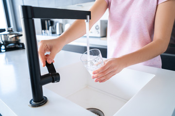 Fototapeta na wymiar Woman pours fresh filtered purified water for drinking from a tap into a glass at kitchen at home. Healthy lifestyle
