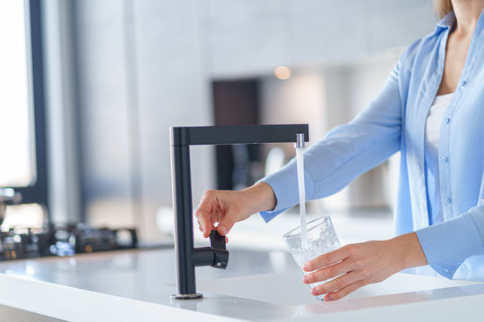 Woman pours fresh filtered purified water from a tap into a glass at kitchen at home for drinking