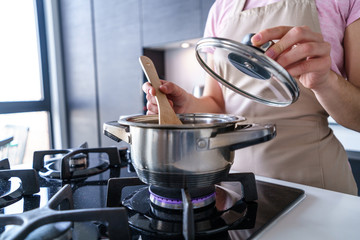 Woman housewife in apron using steel metal saucepan for preparing dinner in the kitchen at home. Kitchenware for cooking food - obrazy, fototapety, plakaty