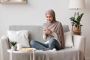 Happy arabic woman in hijab using smartphone on sofa at home