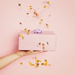Holiday confetti background with gift or present box in woman hand on pink. Beautiful Birthday...