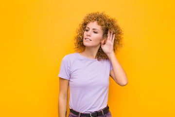 young afro woman looking serious and curious, listening, trying to hear a secret conversation or gossip, eavesdropping against orange wall