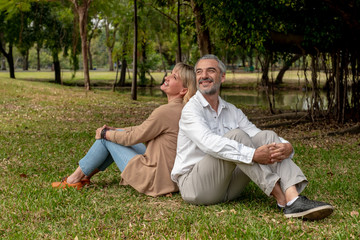 Happy senior Caucasian couple sitting back to back in the park. Smiling senior couple sitting together in the park