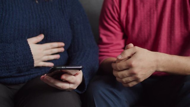 Close-up of deaf diverse people's hands browsing online on smart phone, discussing social media content , communicating with sign language while relaxing on sofa in domestic interior.