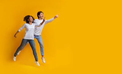 Fototapeta na wymiar Multiracial couple pointing at copy space and jumping over yellow background