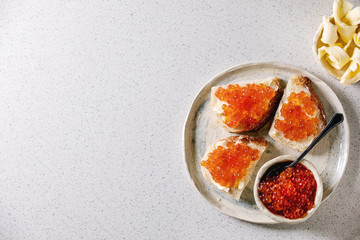 Red salmon caviar in bowl and on wheat bread, served with butter on ceramic plate over grey spotted background. Flat lay, space