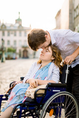 Close-up portrait of happy young woman in wheelchair and her husband kissing her forehead, walking...
