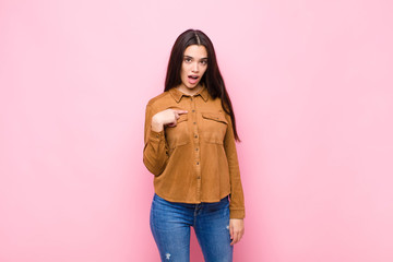 young pretty woman looking shocked and surprised with mouth wide open, pointing to self against...