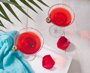 Red exotic alcoholic cocktail in clear glasses and red rose petals on the wooden white table for romantic dinner.