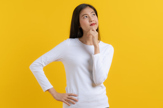 Cute asian young woman in white casual dress looking up and thinking / imagination isolated on yellow background in studio.