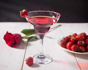 Red exotic alcoholic cocktail in clear glass, plate with fresh strawberries and red rose on the wooden white table for romantic dinner.