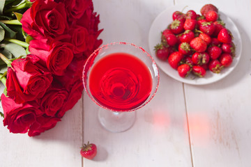 Red exotic alcoholic cocktail in clear glass, plate with fresh strawberries and red roses on the wooden white table for romantic dinner.
