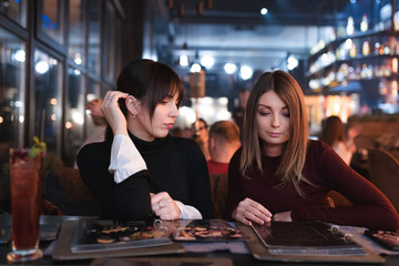 Two pretty woman are studying a menu in sushi bar. One has already chose a cocktail.