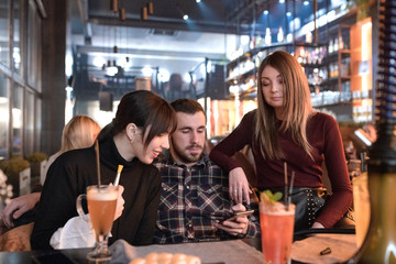 Young hipster friends date in restaurant. Handsome man sitting between two pretty women and they watching in the cellphone device.