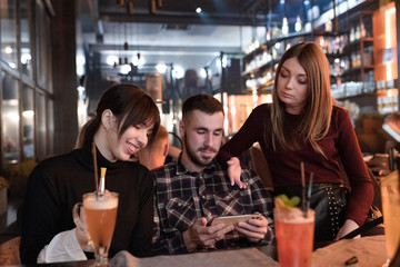 Young hipster friends date in restaurant. Handsome man sitting between two pretty women and they watching in the cellphone device.