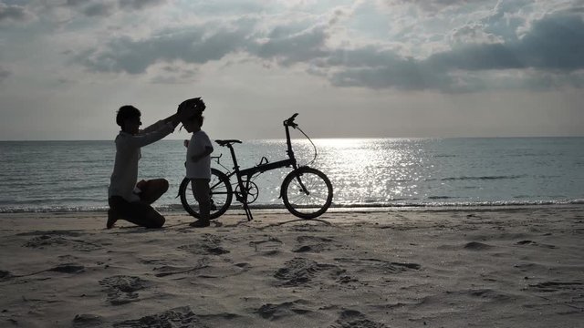 Asian family On Summer Vacation Father wears a hat for his son for safety in riding bicycles on beach. Holiday and Travel concept. Silhouette Slow Motion