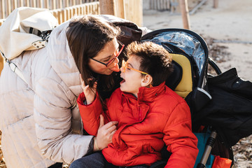 una madre juega con su hijo con pluridiscapacidad en un parque, el niño va con silla de ruedas, es...