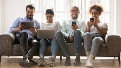 Smiling diverse young people sit on couch using gadgets