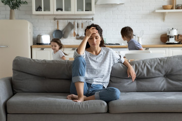 Exhausted mother sitting on couch while kids running at home - obrazy, fototapety, plakaty