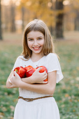 Cute girl in white dress holding red apples. Beautiful Girl Eating Organic Apple in the Orchard. Harvest Concept. Garden, Toddler eating fruits at fall harvest. Apple pie