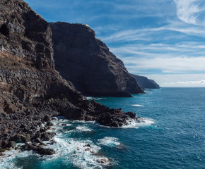 Fototapeta na wymiar High steep Volcanic rock cliffs of black lava on the rocky shore with crushing white waves over the Atlantic Ocean. Blue sky background. La Palma, Canary Islands.