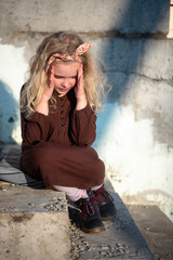 girl blond curls holding on to her head sitting on the steps in a dress and boots on a sunny day