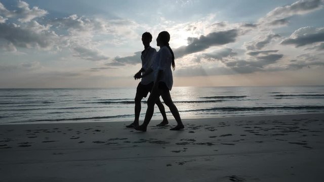 Side view Happy Asian young couple in love holding hands walking and play together on the beach. Background sunset in sea. Holiday rest and Travel concept. Slow Motion