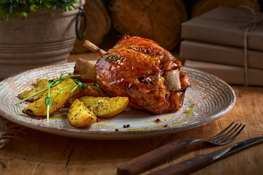 Pork knuckle with baked potatoes on rustic kitchen table at dark wooden background, front view. Pork leg done, German food