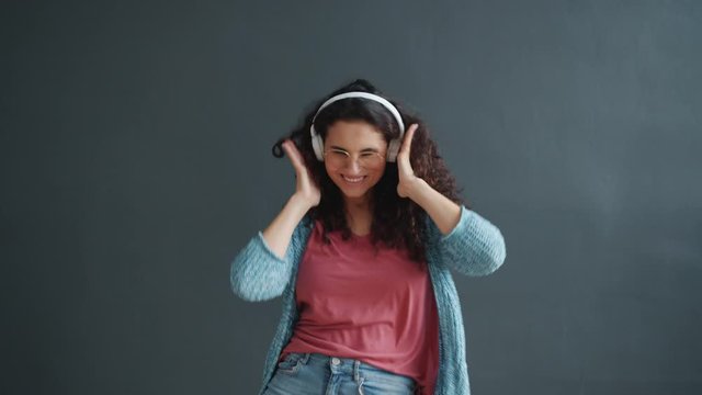Joyful mixed race girl wearing wireless headphones is dancing on dark gray background having fun enjoying song alone. Modern technology and people concept.