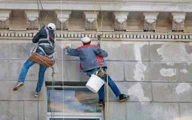 Construction workers hanging on rope and painting building facade, working with finishing trowel at...