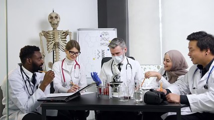 Four young confident multiracial male and female future medical workers carefully listening the respected bearded doctor-mentor with gray hair during joint work