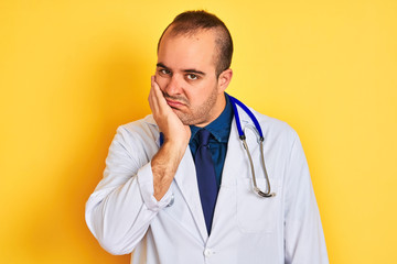 Young doctor man wearing coat and stethoscope standing over isolated yellow background thinking looking tired and bored with depression problems with crossed arms.