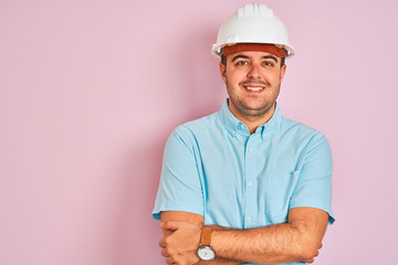 Young architect man wearing security helmet standing over isolated pink background happy face smiling with crossed arms looking at the camera. Positive person.