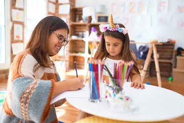 Beautiful teacher and toddler wearing unicorn diadem drawing using paper and pencils around lots of...