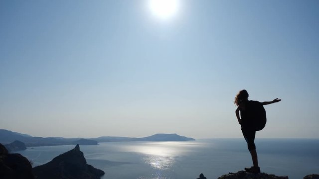 The girl conquered a tall rock and admires the view of the sea and the salt in the clear sky. Athletic woman raises her hands and enjoys the beauty of nature under the rays of the sun in slow motion.