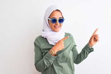Young Arab woman wearing hijab and summer sunglasses over isolated background smiling and looking at the camera pointing with two hands and fingers to the side.