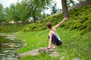 Naklejka na ściany i meble A young sports girl practices yoga on a green lawn by the river, yoga assans posture. Meditation and unity with nature