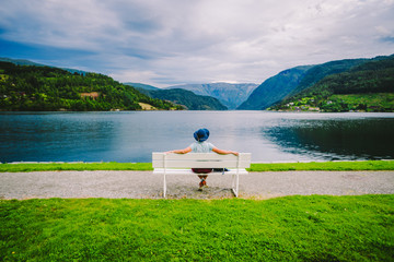 Woman sitting on a bench looking at the fjord in Ulvik, Norway. Fjord coastal promenade in Ulvik, Hordaland county, Norge. Lonely tourist in hat sits back on bench and admires scenery Scandinavia