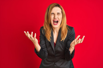 Young caucasian business woman wearing a suit over isolated red background crazy and mad shouting and yelling with aggressive expression and arms raised. Frustration concept.