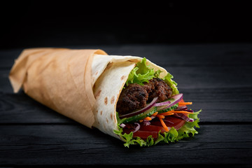 Shawarma rolled in lavash, moist grilled meat with onion, herbs and vegetables on wooden black background.