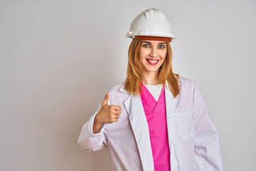 Redhead caucasian woman engineer wearing safety helmet over isolated background doing happy thumbs up gesture with hand. Approving expression looking at the camera with showing success.