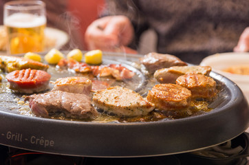 pieces of meat are roasting on a gourmet set. Smoke is coming from the hot plate. Beer and hand of person in background.