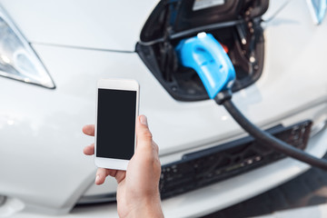 Transportation. Young man traveling by electric car stop at chraging station looking at smartphone screen copy space close-up while having vehicle fully charged