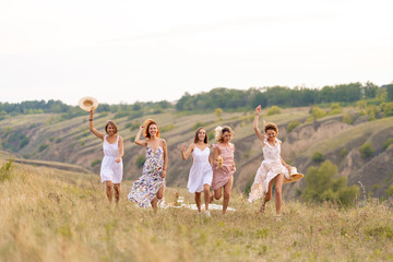 Fototapeta premium The company of cheerful female friends have a great time together on a picnic in a picturesque place overlooking the green hills. Girls in white dresses dancing in the field