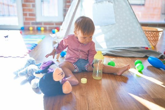 Adorable toddler holding feeding bottle around lots of toys at kindergarten