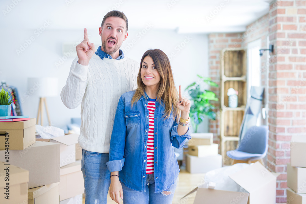 Sticker young beautiful couple standing at new home around cardboard boxes pointing finger up with successfu