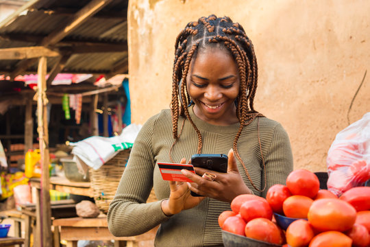 Young African Woman Selling In A Local African Market Using Her Mobile Phone And Credit Card To Do A Transaction Online