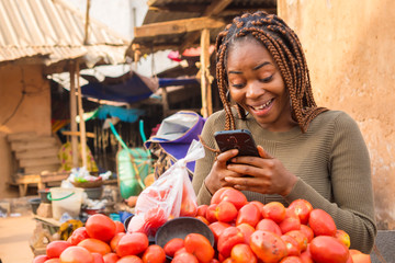 beautiful young african woman in a local african market viewing content on her phone looking...
